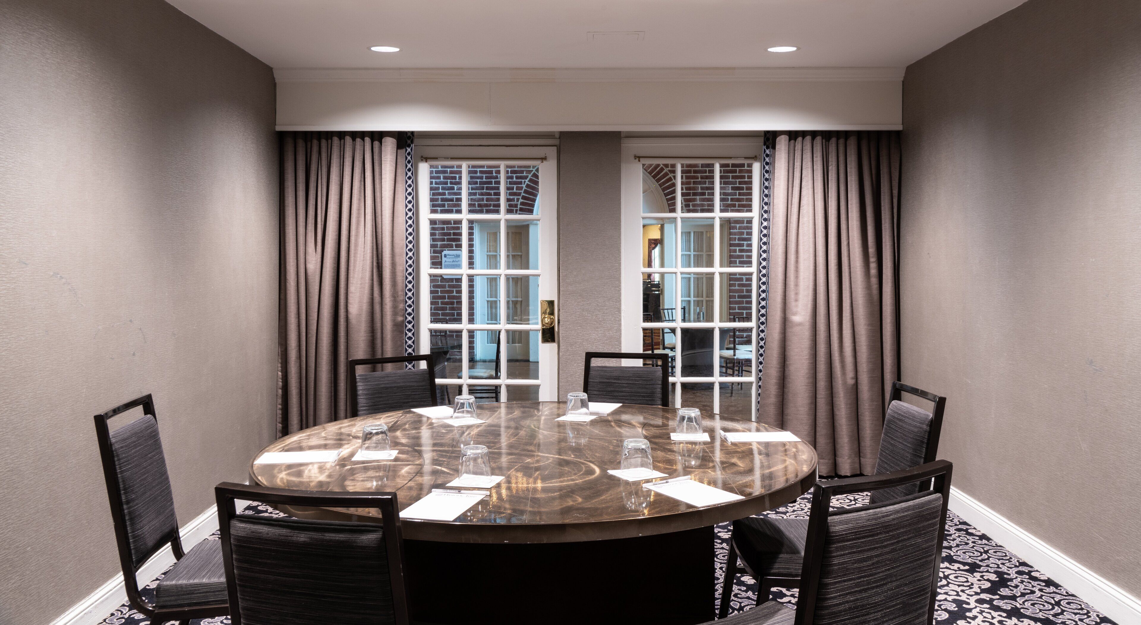 A conference room with a round table, six chairs, and a view through large windows with curtains.