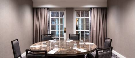 A conference room with a round table, six chairs, and a view through large windows with curtains.