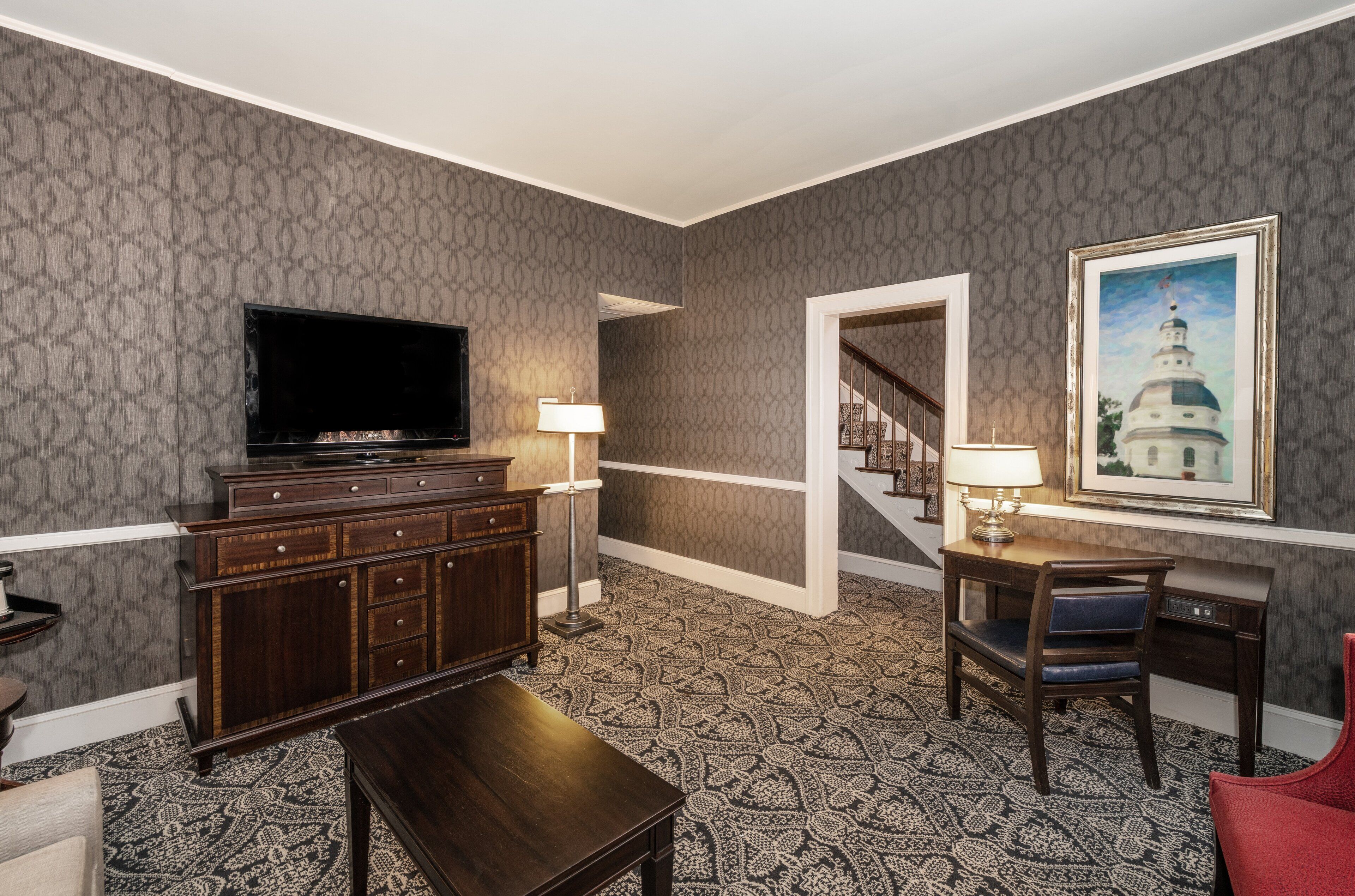 A hotel room with a large flat-screen TV, a wooden cabinet, a desk with a chair, a patterned carpet, and a framed picture on the wall.