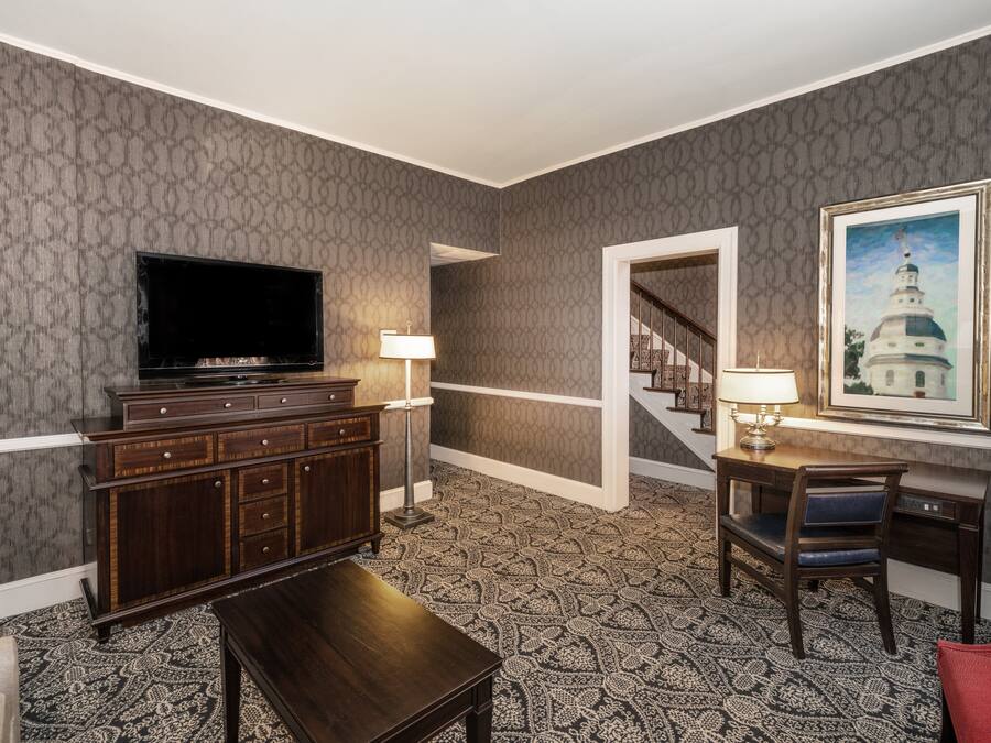 A hotel room with a large flat-screen TV, a wooden cabinet, a desk with a chair, a patterned carpet, and a framed picture on the wall.