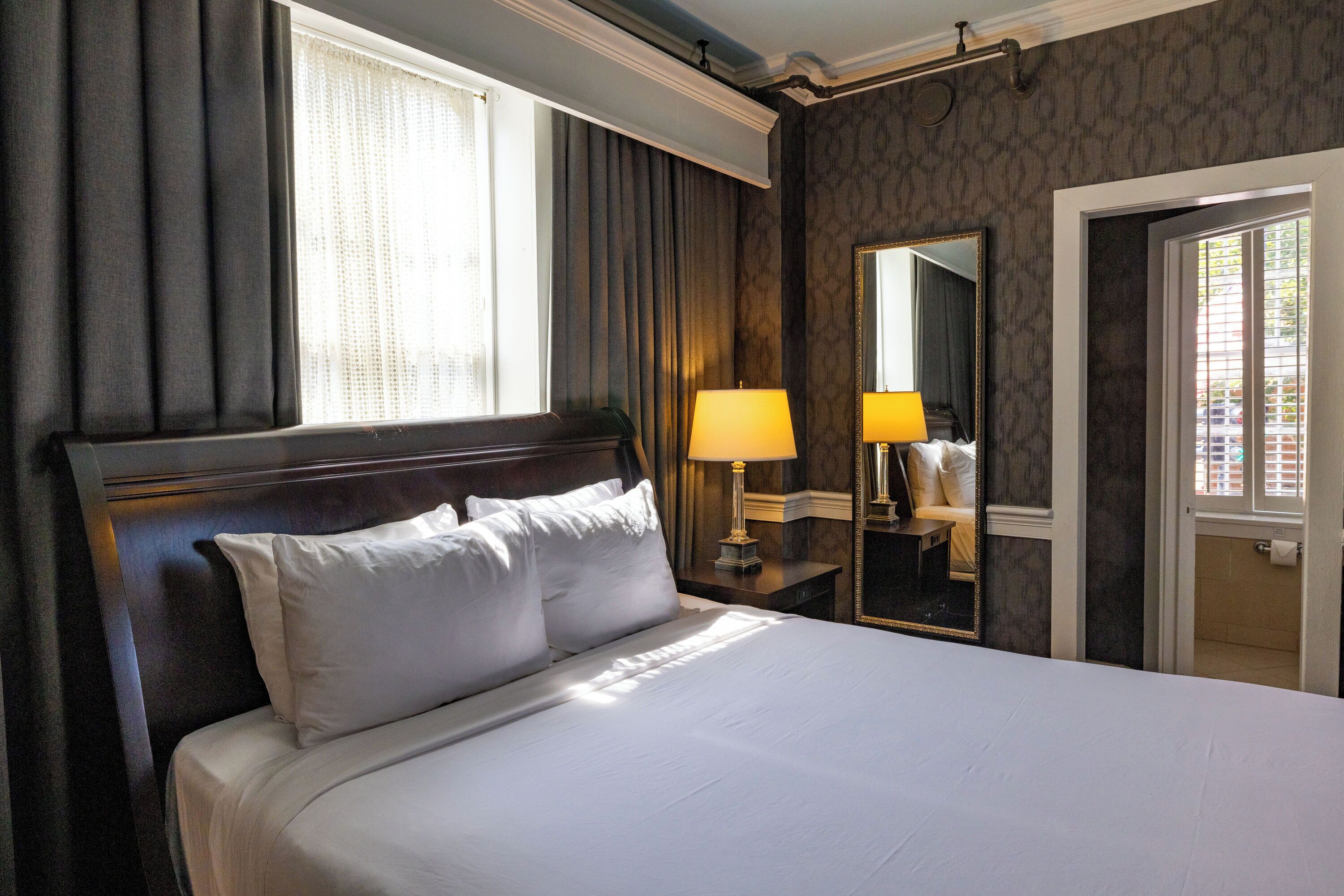A hotel room with a large bed, a nightstand with a lamp, a mirror, and a view of the outside through a window.
