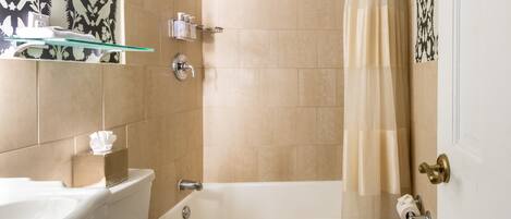 Un baño con bañera, inodoro y un estante de vidrio con artículos de tocador.