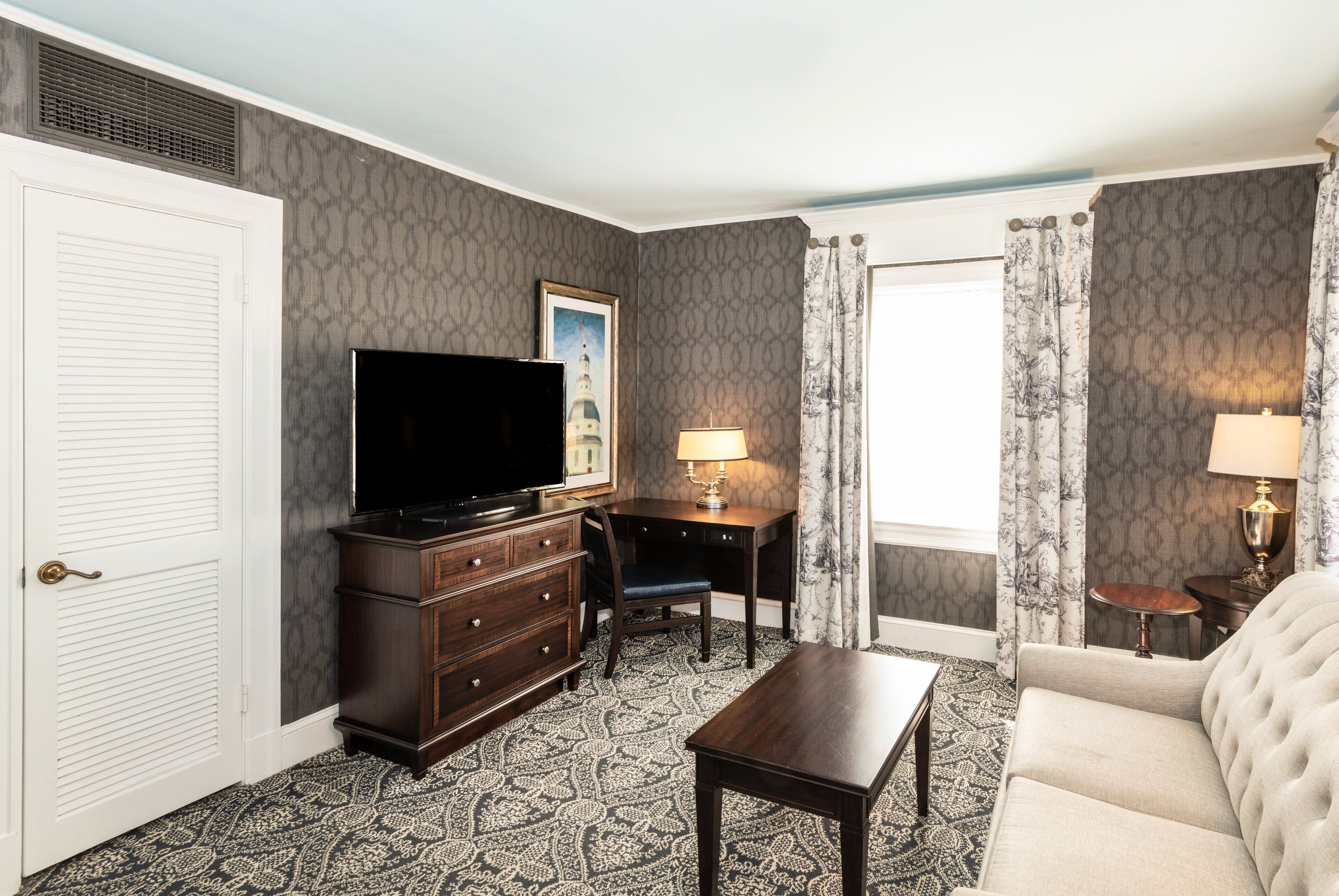 Hotelkamer met flatscreen televisie, bureau, stoel, bank, klein tafeltje en een kamerbreed tapijt met dessin.