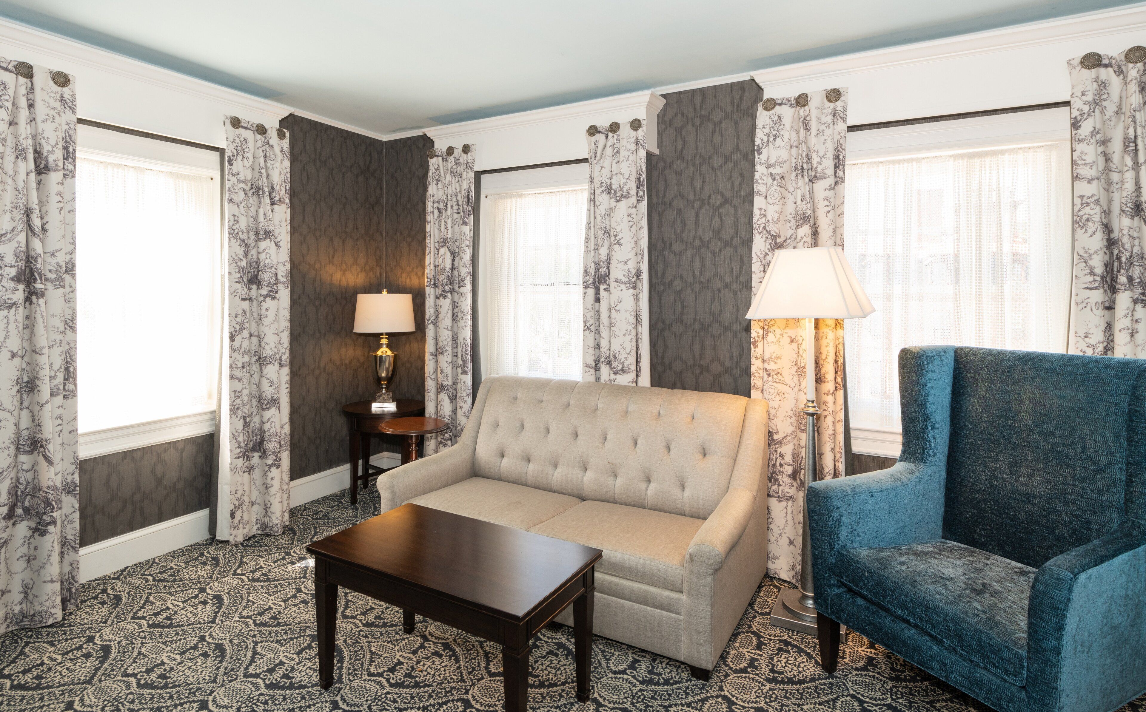 Una habitación con sofá beige, sillón azul, una mesita y una lámpara. La habitación tiene cortinas y alfombra estampadas.