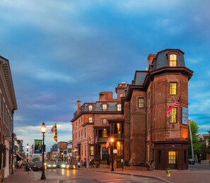 A historic brick building with a corner turret and multiple windows, located on a city street with flags and street lamps.