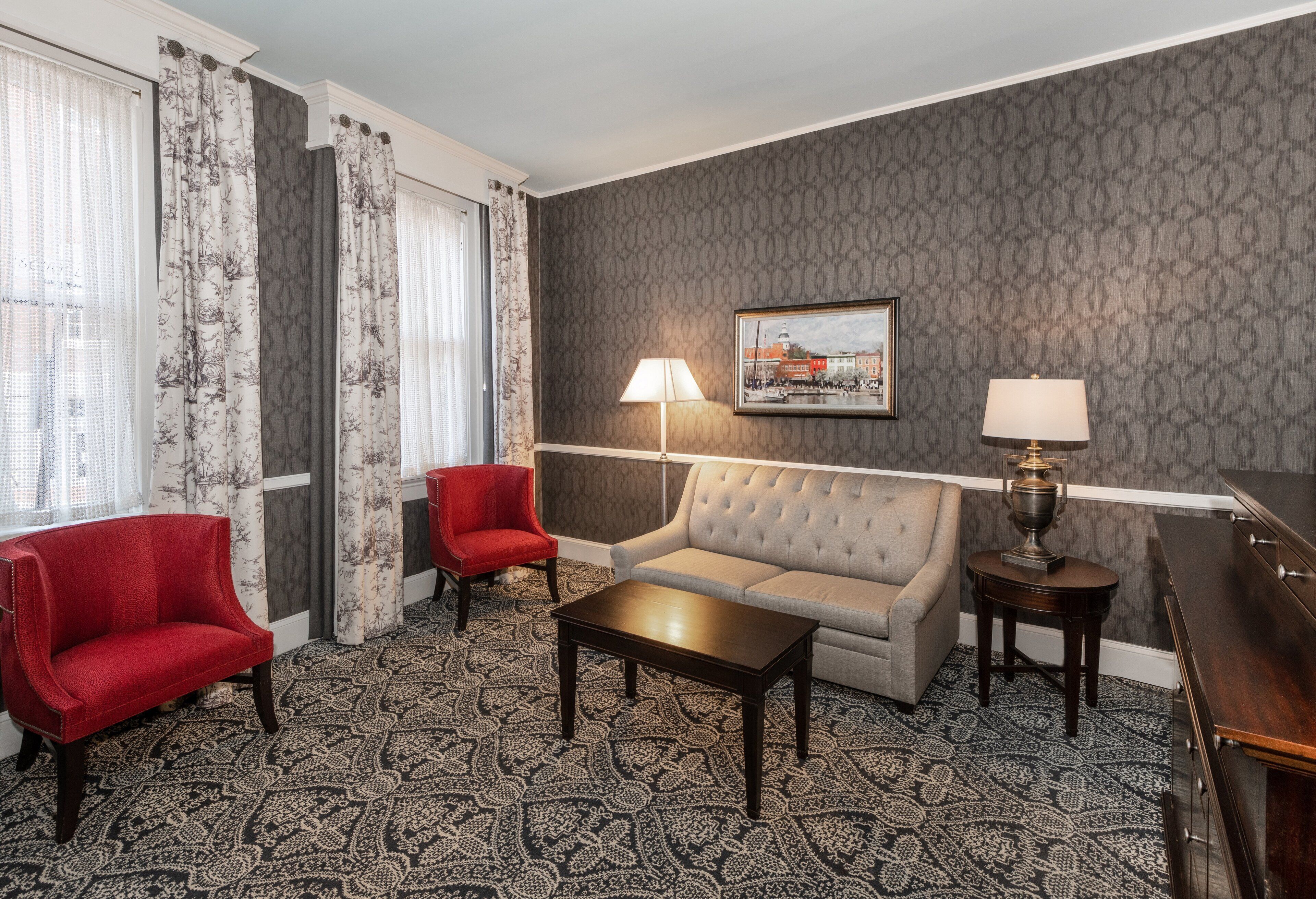 A hotel room with a grey patterned wall, a grey sofa, a red armchair, a wooden coffee table, two lamps, and a framed picture on the wall.