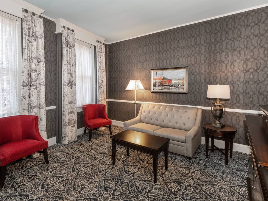 A hotel room with a grey patterned wall, a grey sofa, a red armchair, a wooden coffee table, two lamps, and a framed picture on the wall.