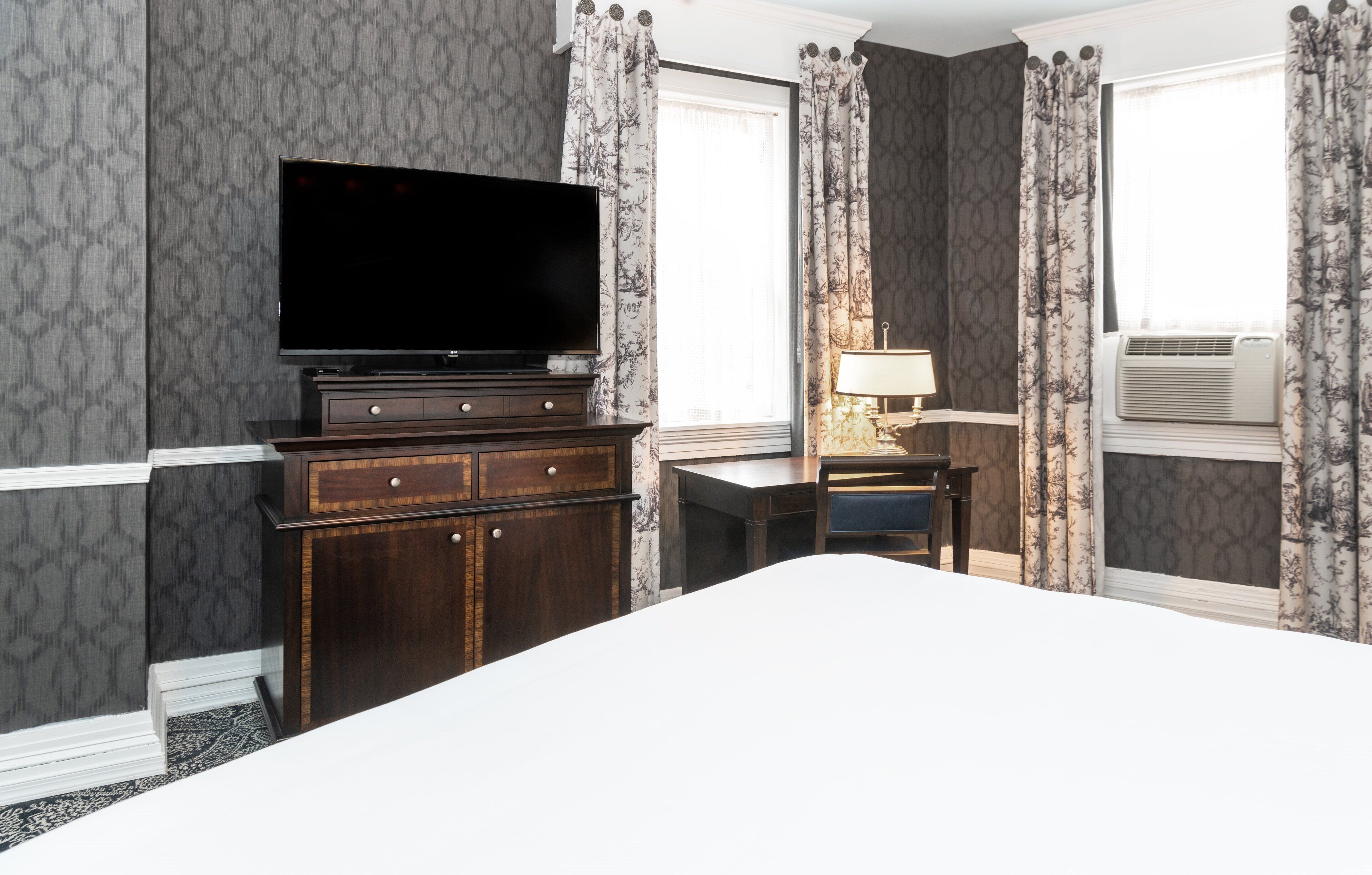 Une chambre d’hôtel avec un grand lit, un téléviseur à écran plat posé sur une commode en bois, un bureau avec une lampe et un appareil de climatisation.
