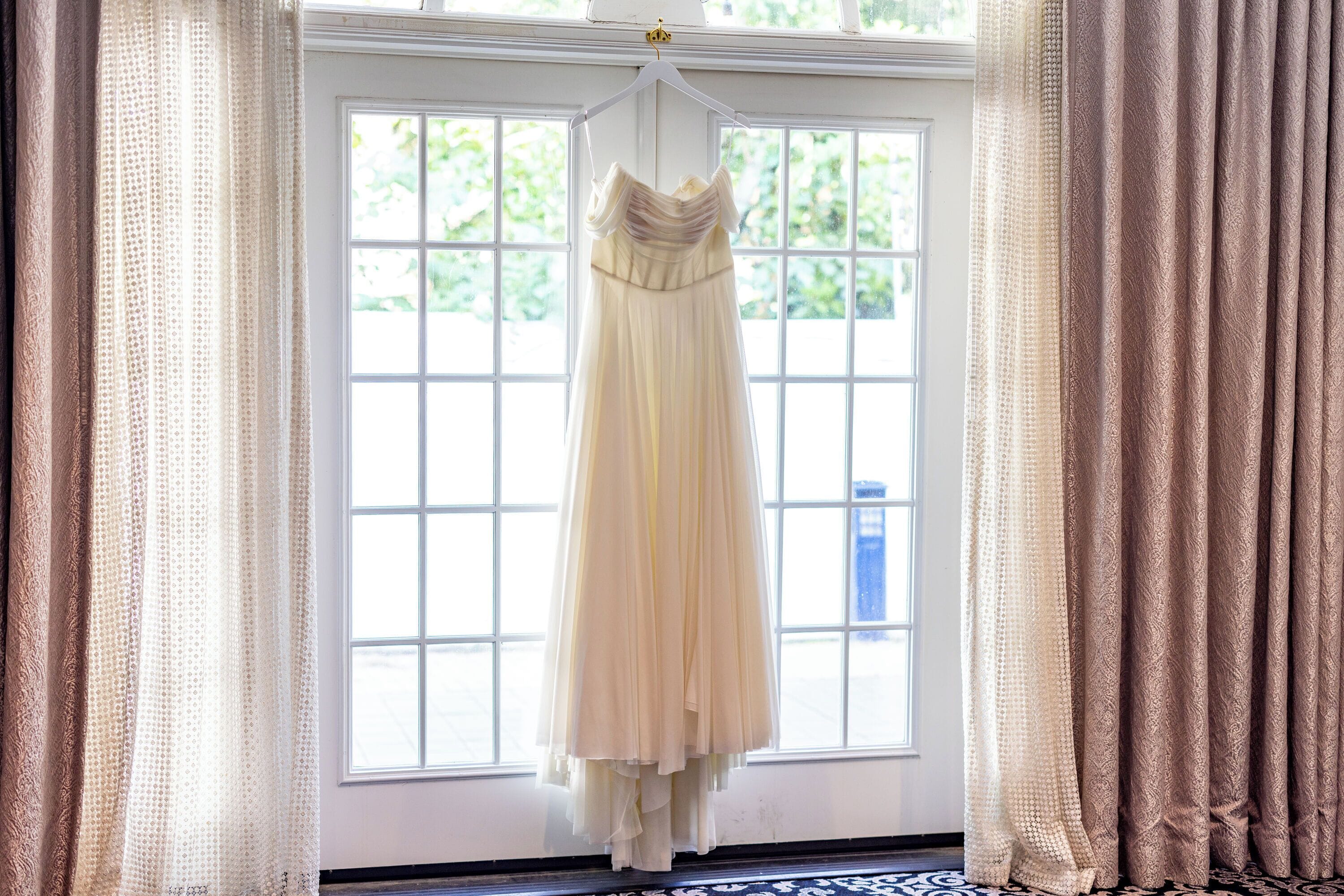 A wedding dress hanging in front of a window with a view of greenery outside.