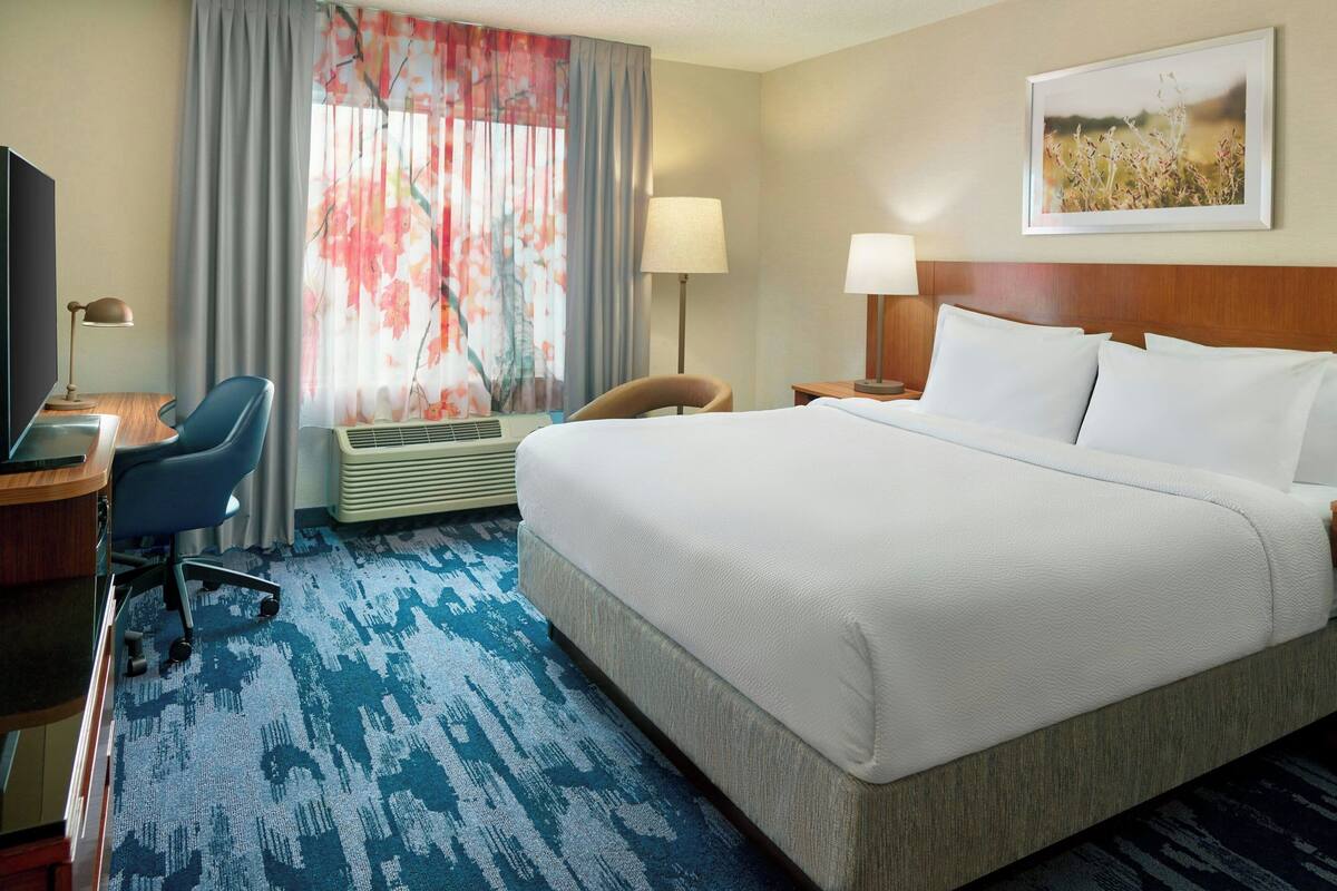 A hotel room with a large bed, a desk with a chair, a lamp, and a window with curtains.