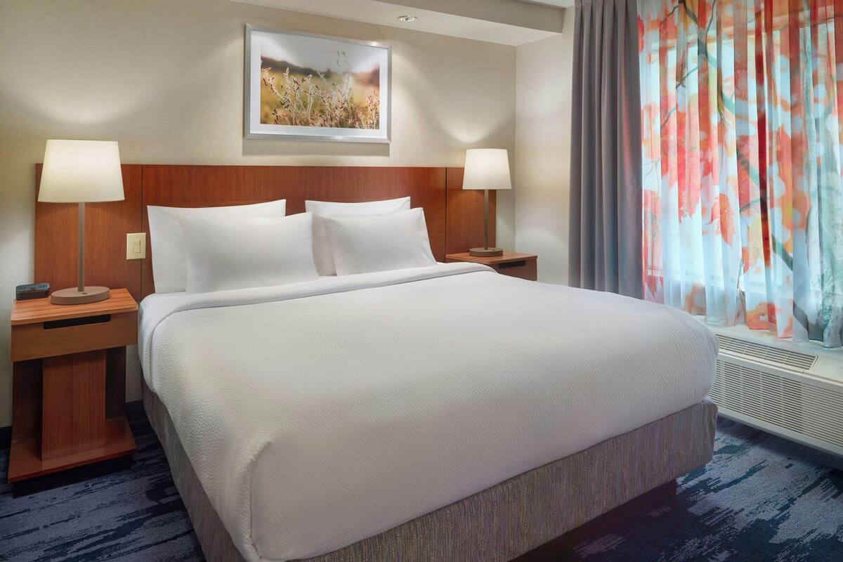A large bed with white linens, two bedside tables with lamps, a framed picture above the bed, and a window with colorful curtains.