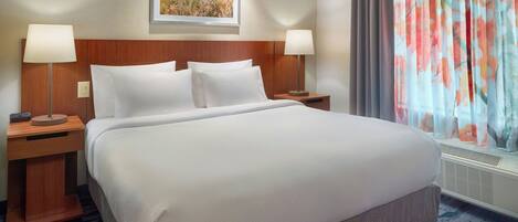 Una cama grande con ropa blanca, dos mesitas de noche con lámparas, un cuadro enmarcado sobre la cama y una ventana con cortinas de colores.