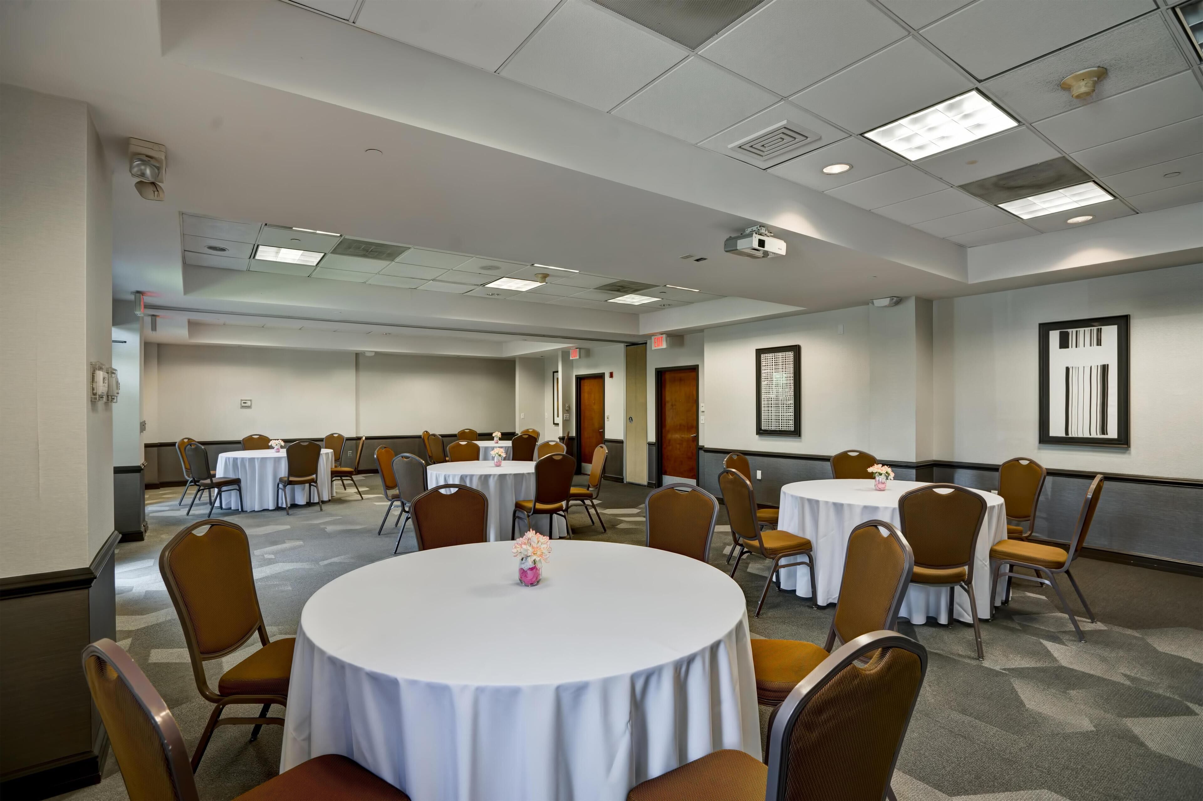 A conference room with multiple round tables and chairs, equipped with ceiling-mounted projectors and recessed lighting.