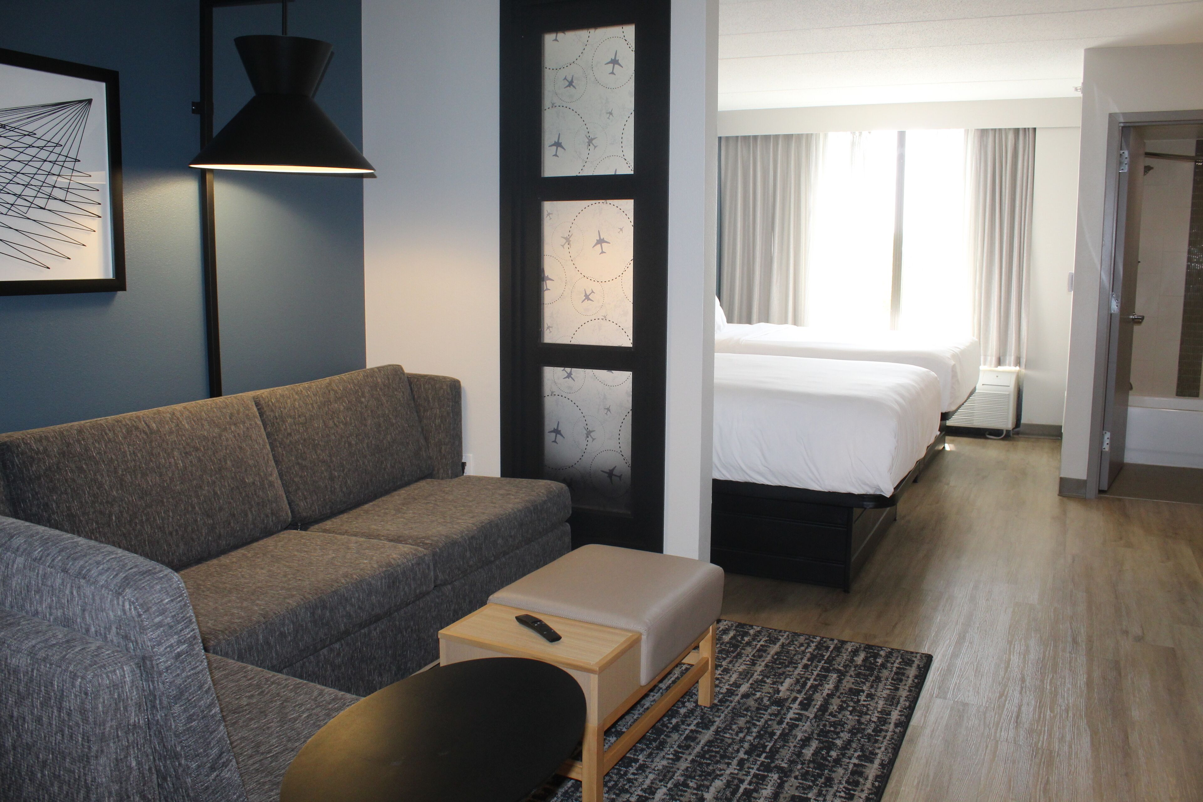 Habitación de hotel con sofá gris, una pequeña mesa de madera con un control remoto, una alfombra estampada, una cama con sábanas blancas y un ventanal con cortinas.
