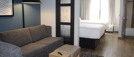 Habitación de hotel con sofá gris, una pequeña mesa de madera con un control remoto, una alfombra estampada, una cama con sábanas blancas y un ventanal con cortinas.