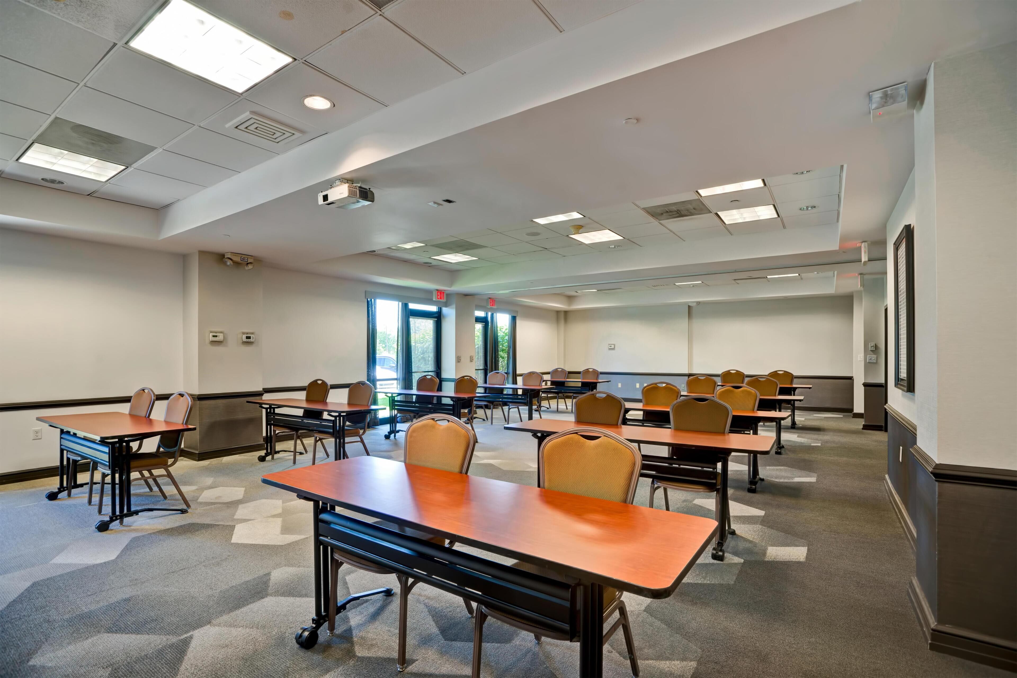 A conference room with multiple tables and chairs, large windows, and a projector on the ceiling.