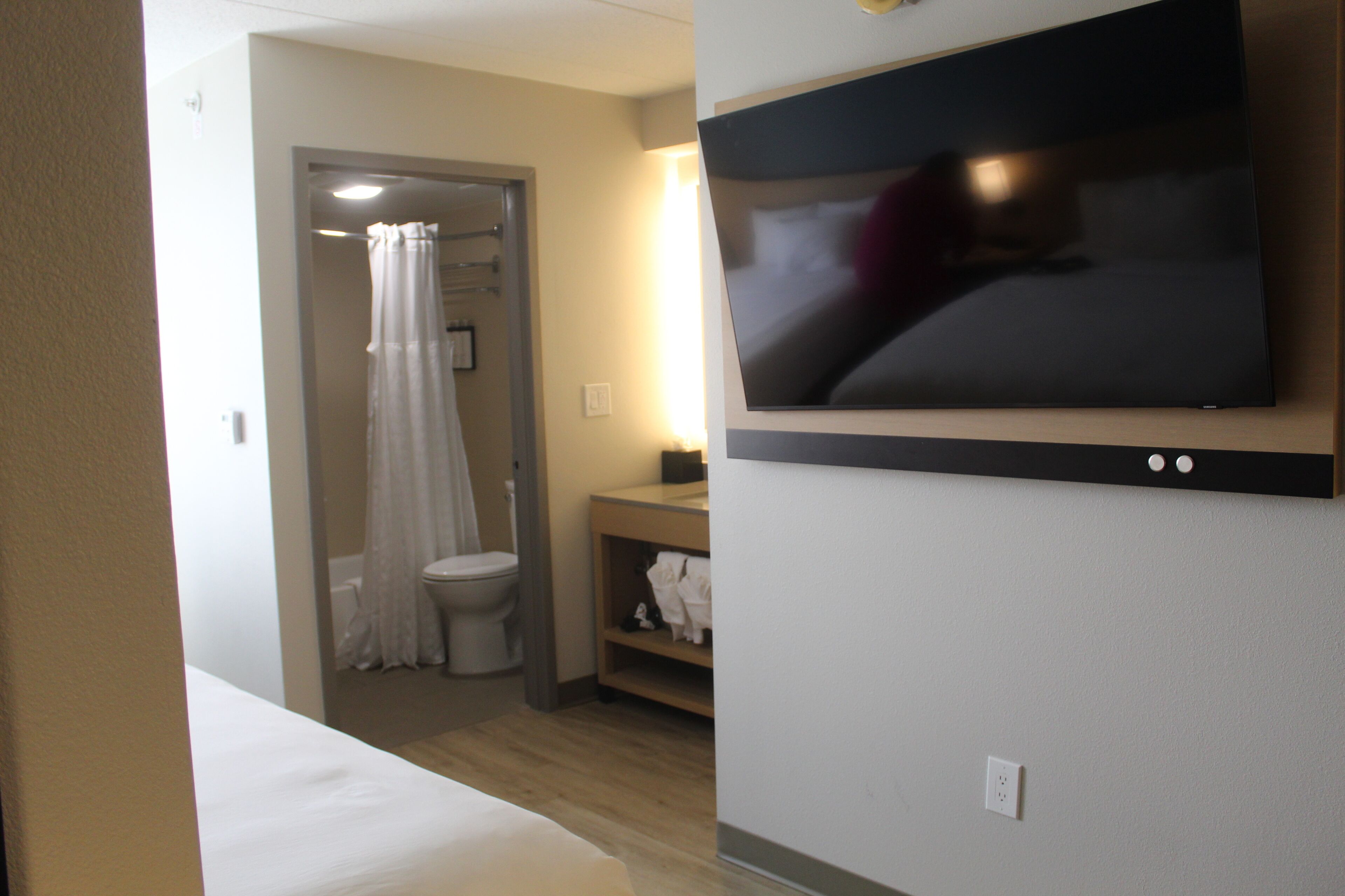 Habitación de hotel con una cama, un televisor montado en la pared, zona de ducha con cortina y un estante con toallas.