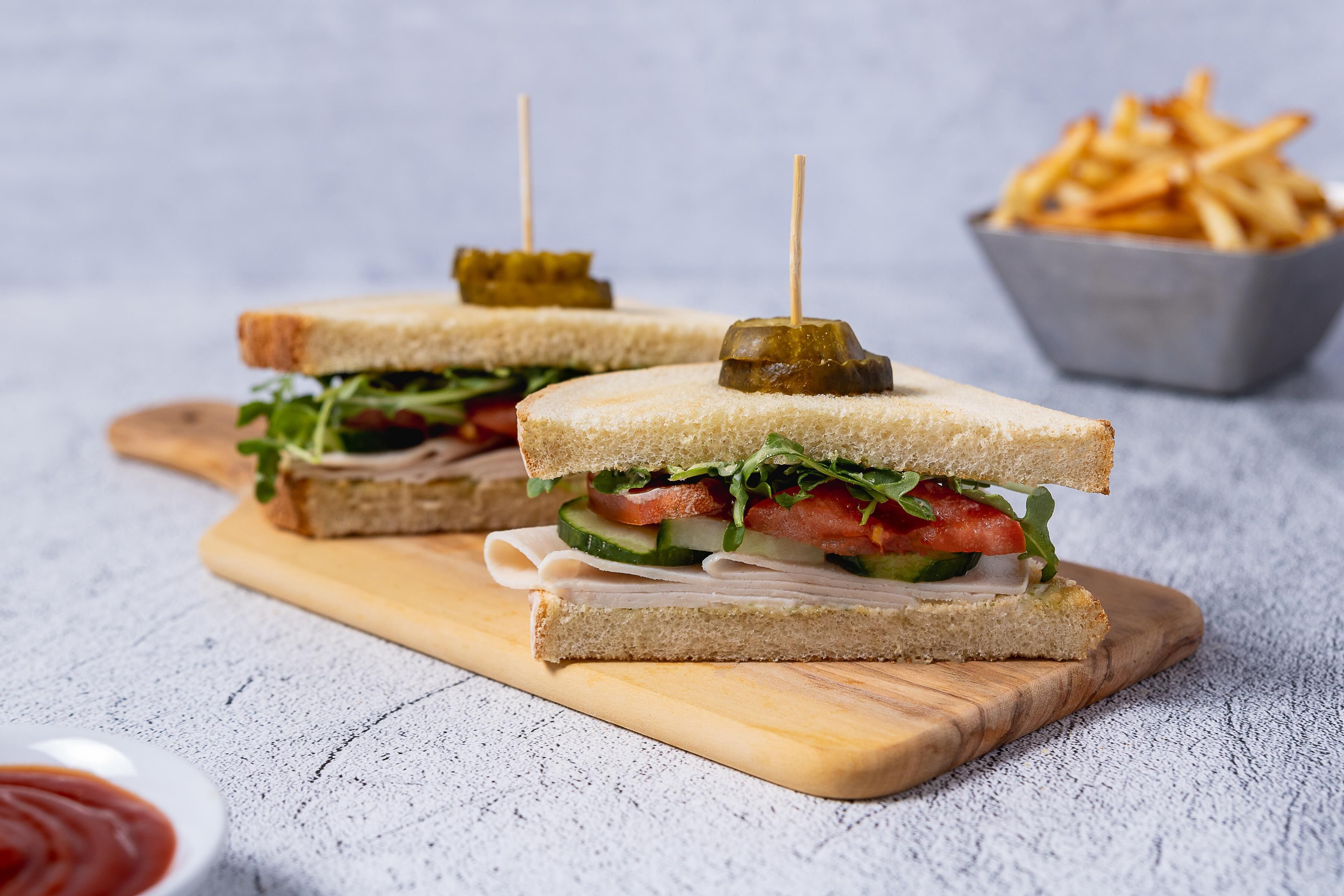 Two sandwiches with toothpicks on a wooden board, a bowl of fries, and a small bowl of ketchup.