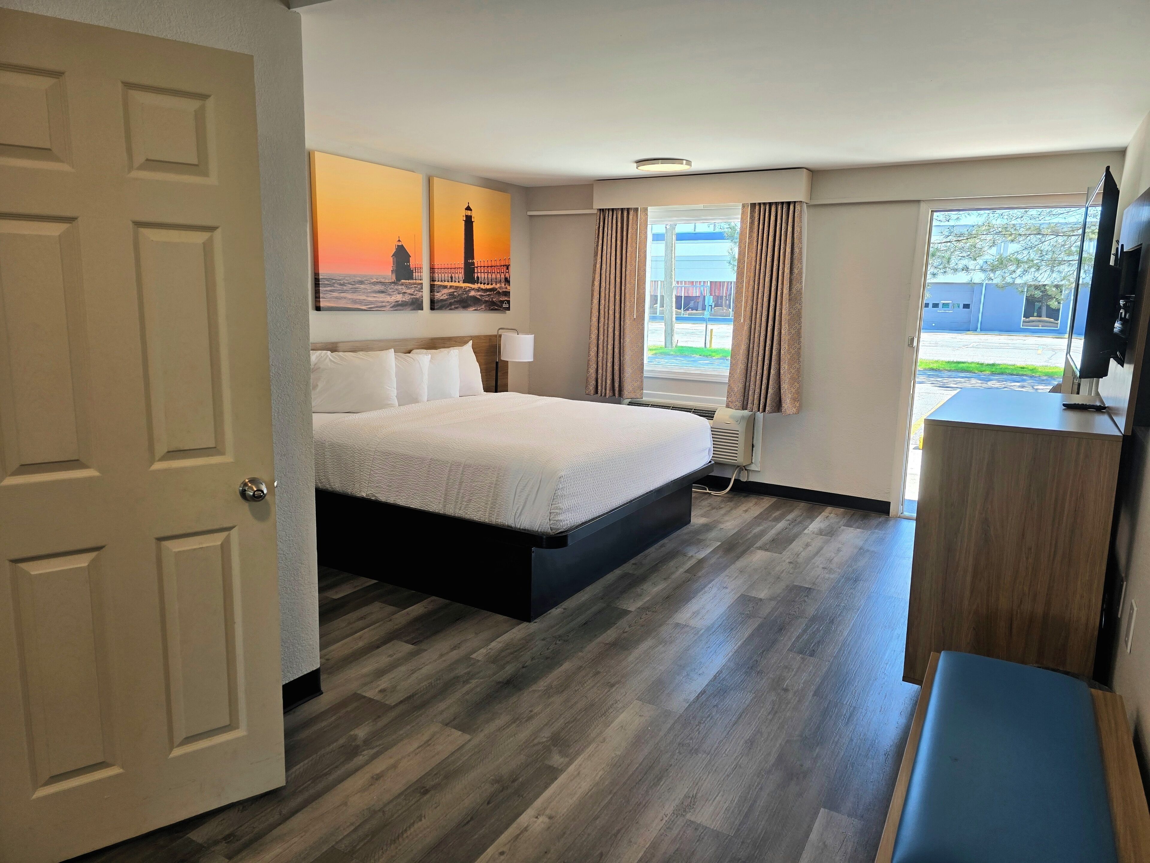 A hotel room with a bed, a desk, a TV, a window with curtains, and a painting of a lighthouse.