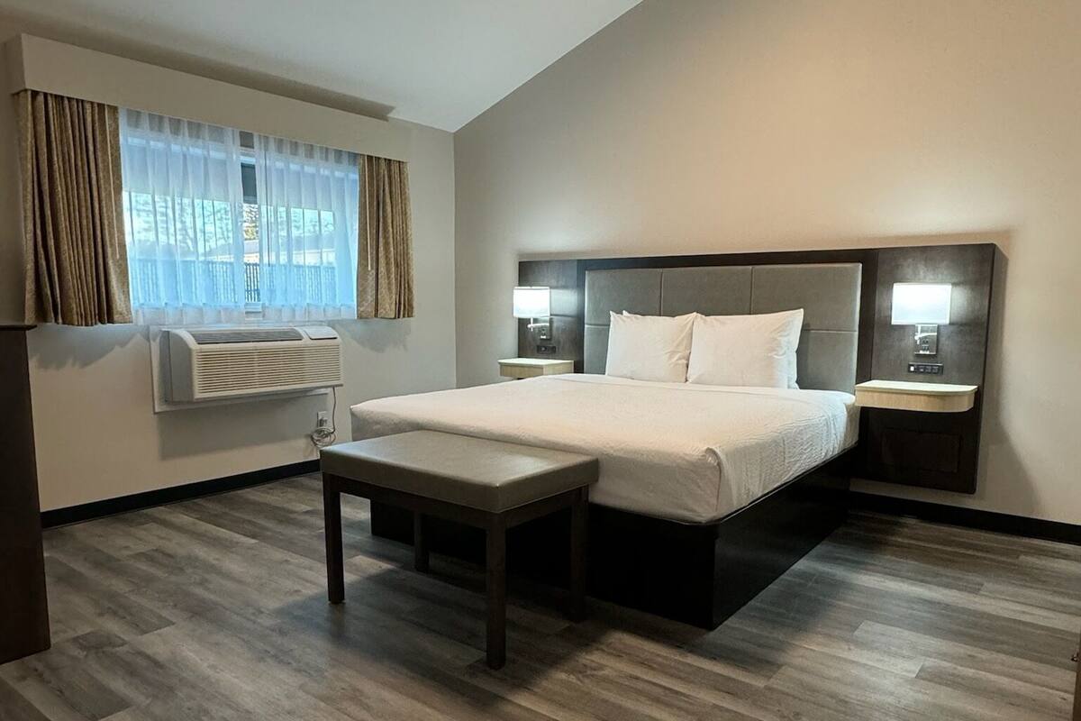 A hotel room with a large bed, bedside tables, a bench, a window with curtains, and an air conditioning unit.