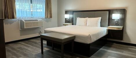 A hotel room with a large bed, bedside tables, a bench, a window with curtains, and an air conditioning unit.