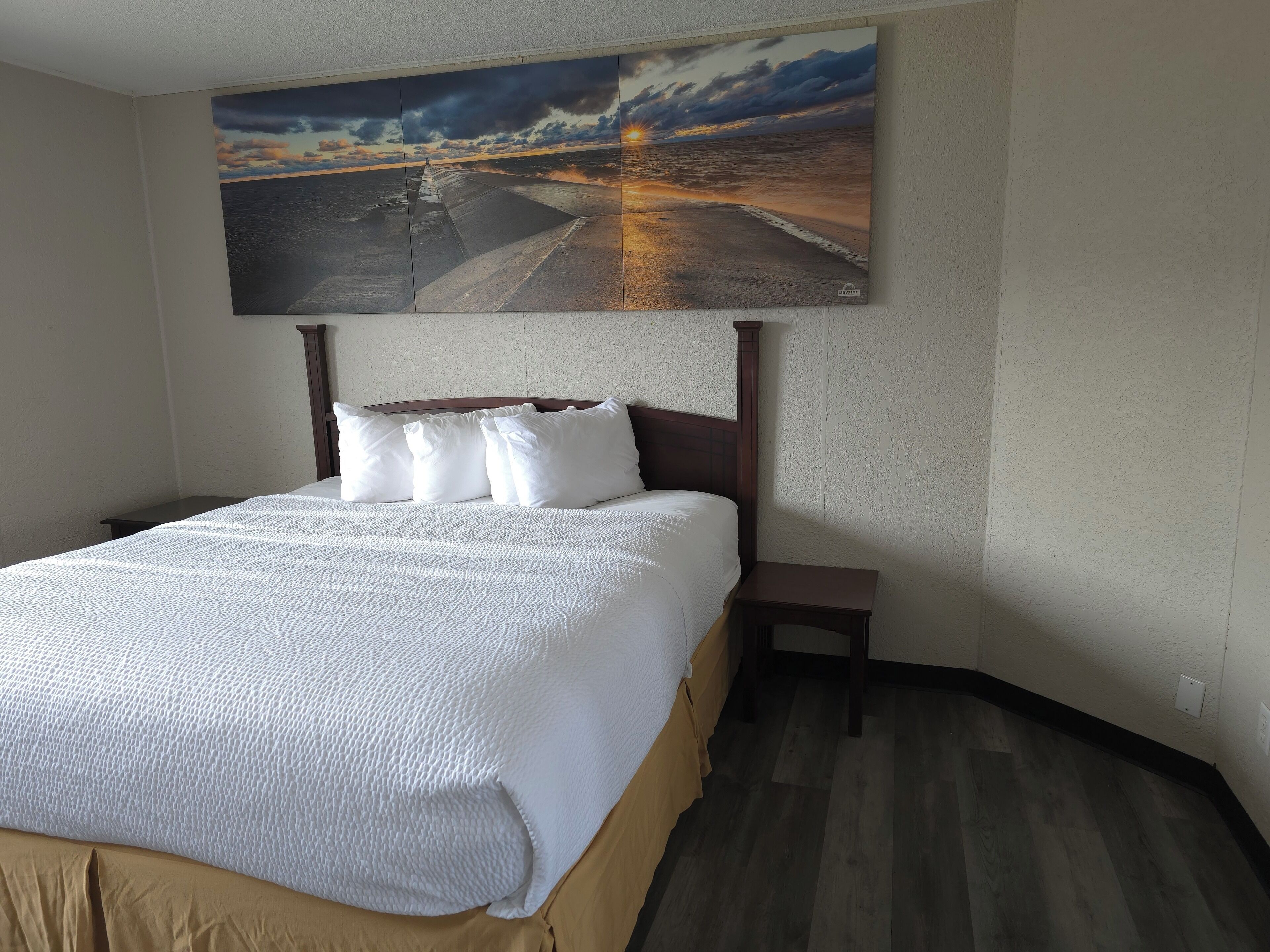 Une chambre d’hôtel avec un grand lit, une tête de lit en bois, une table de chevet et un tableau représentant un coucher de soleil sur l’océan.