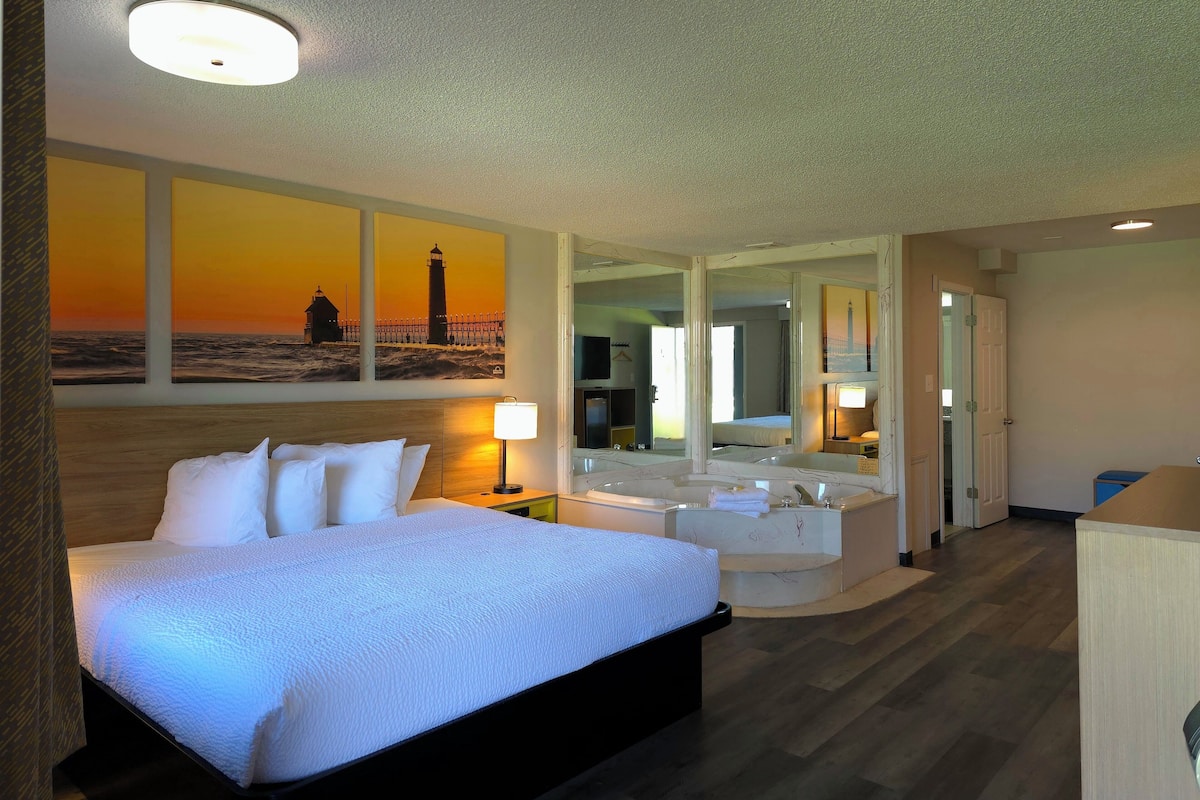 A hotel room with a large bed, a sitting area with a view of the sea, and a bathroom with a bathtub and mirror.
