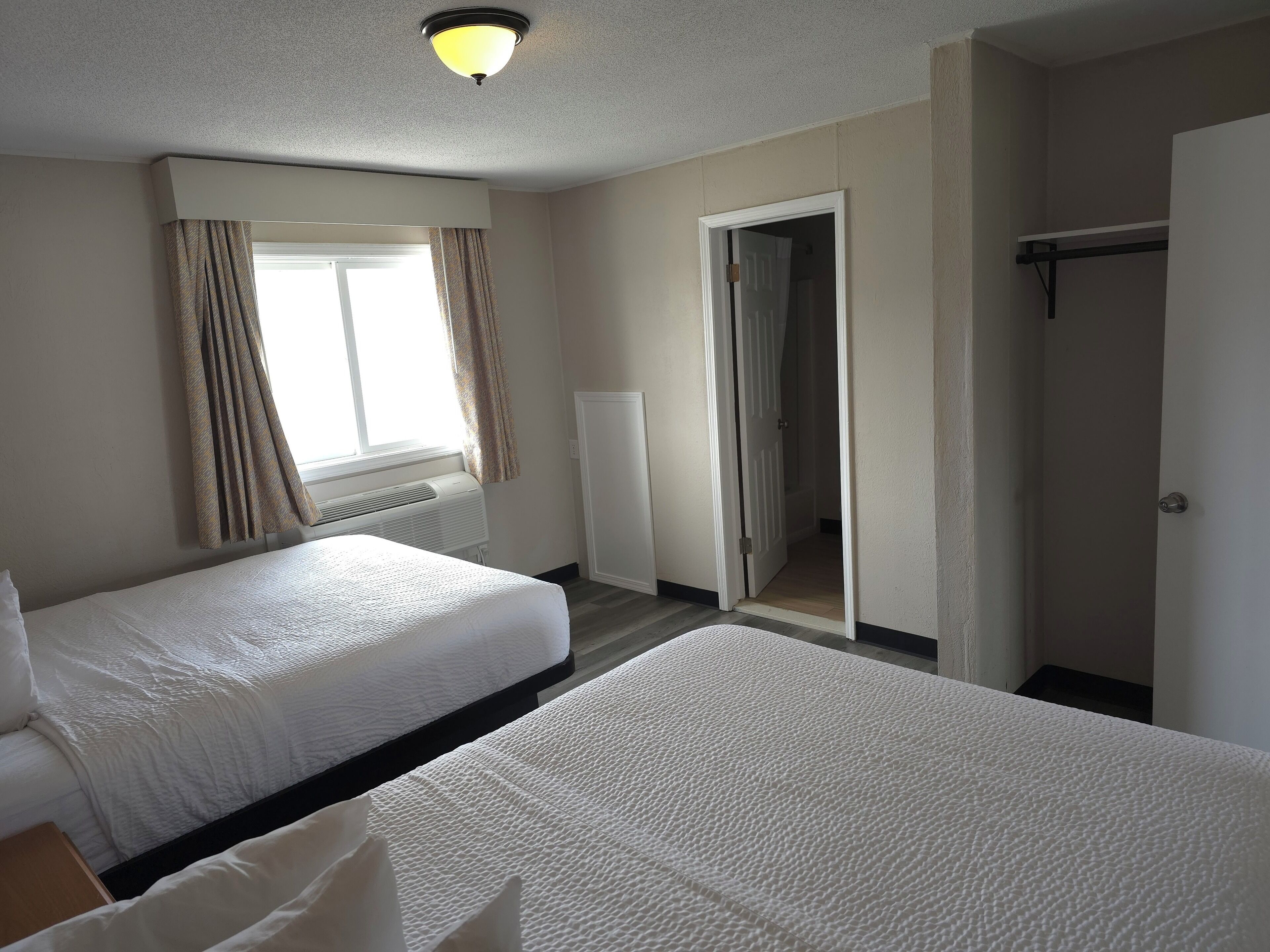 A hotel room with two beds, a window with curtains, a door leading to a hallway, and a ceiling light.