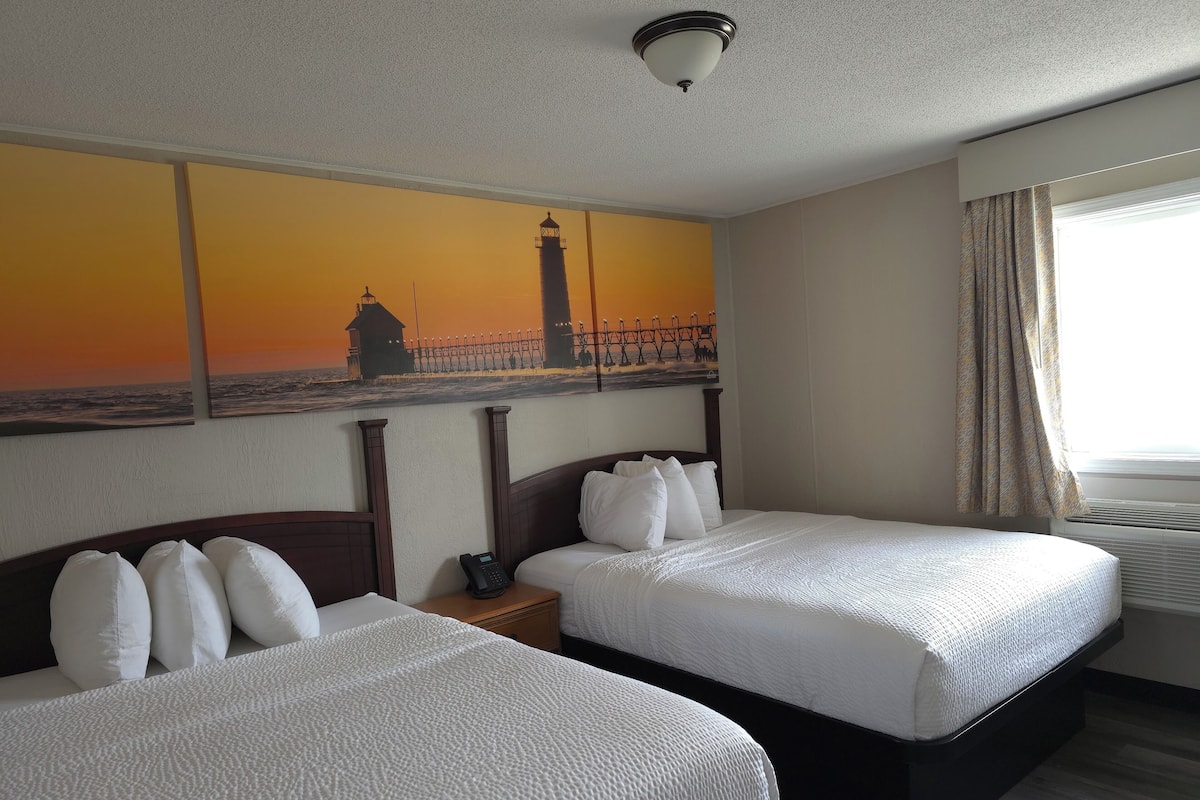A hotel room with two beds, a painting of a lighthouse, a telephone on the nightstand, and a window with curtains.
