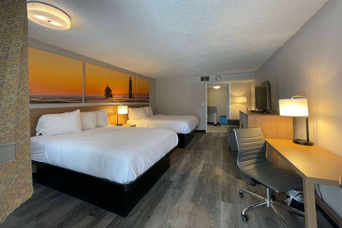 A hotel room with two beds, a desk, a chair, and a large painting of a sunset over the ocean.