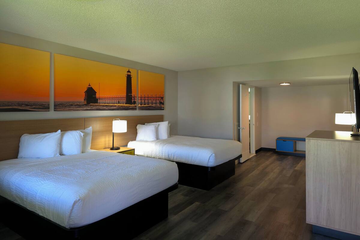 A hotel room with two beds, a painting of a lighthouse, a desk with a lamp, and a TV.