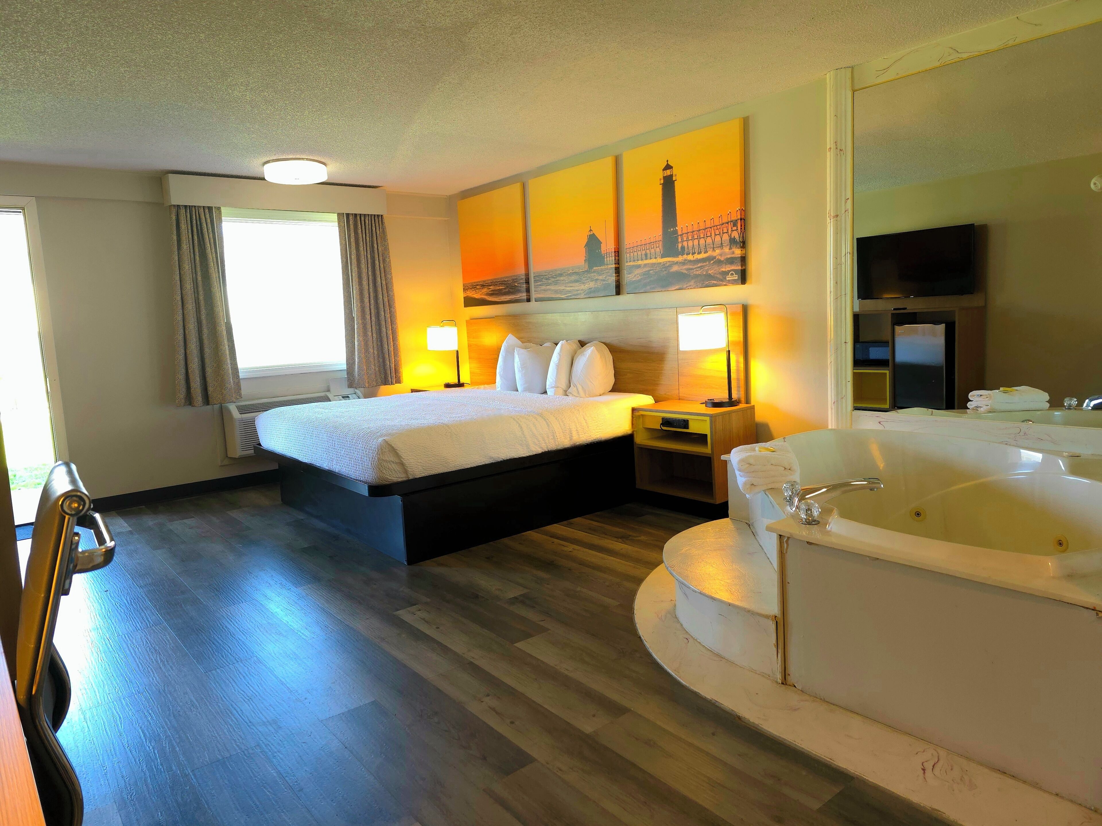 A hotel room with a bed, a large window with curtains, a painting of a lighthouse, a TV, a minibar, and a bathtub.