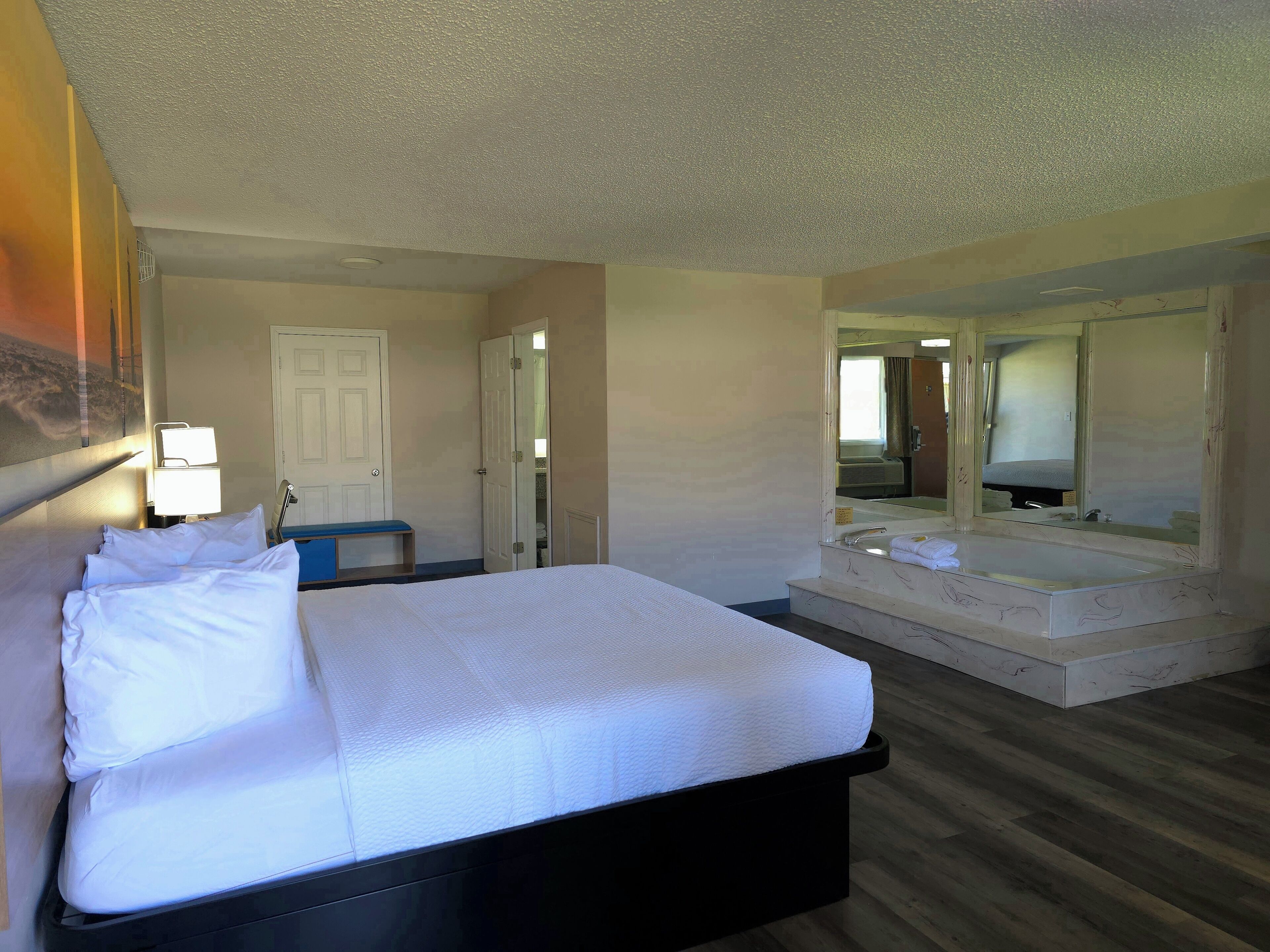 A hotel room with a large bed, a sitting area with a bench, a bathroom with a large mirror, and a door leading to another room.