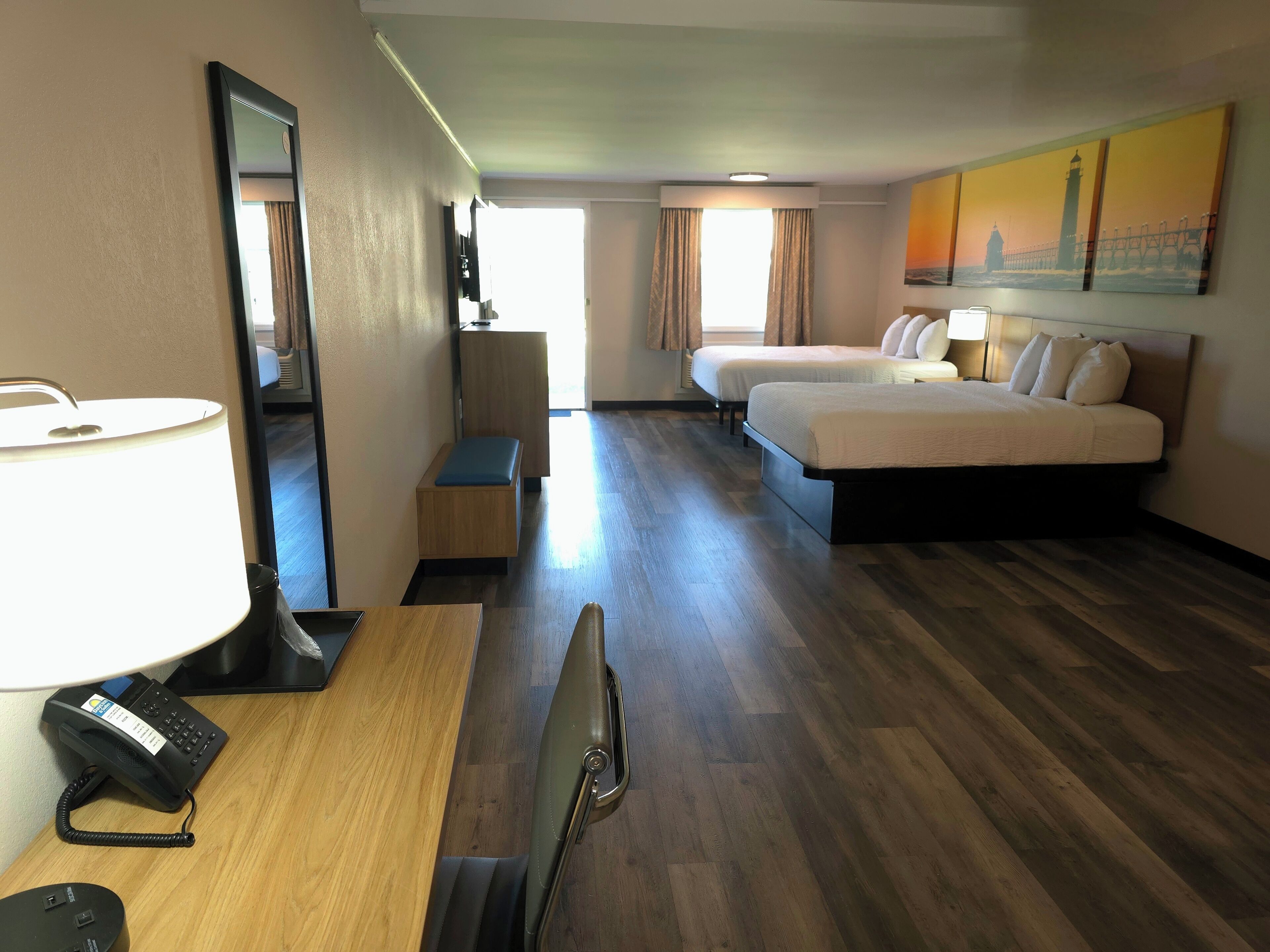 A hotel room with a desk, two beds, a mirror, a lamp, and a painting on the wall.