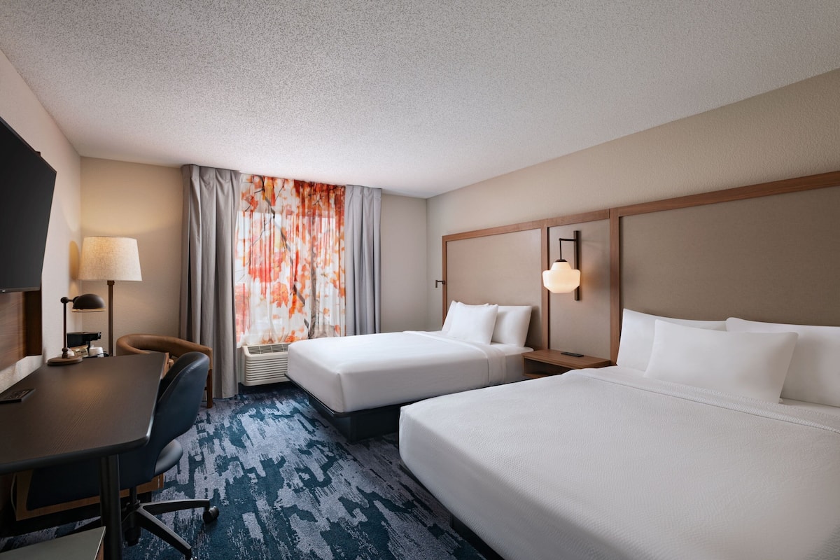 A hotel room with two beds, a desk, a chair, a TV, and a window with curtains.