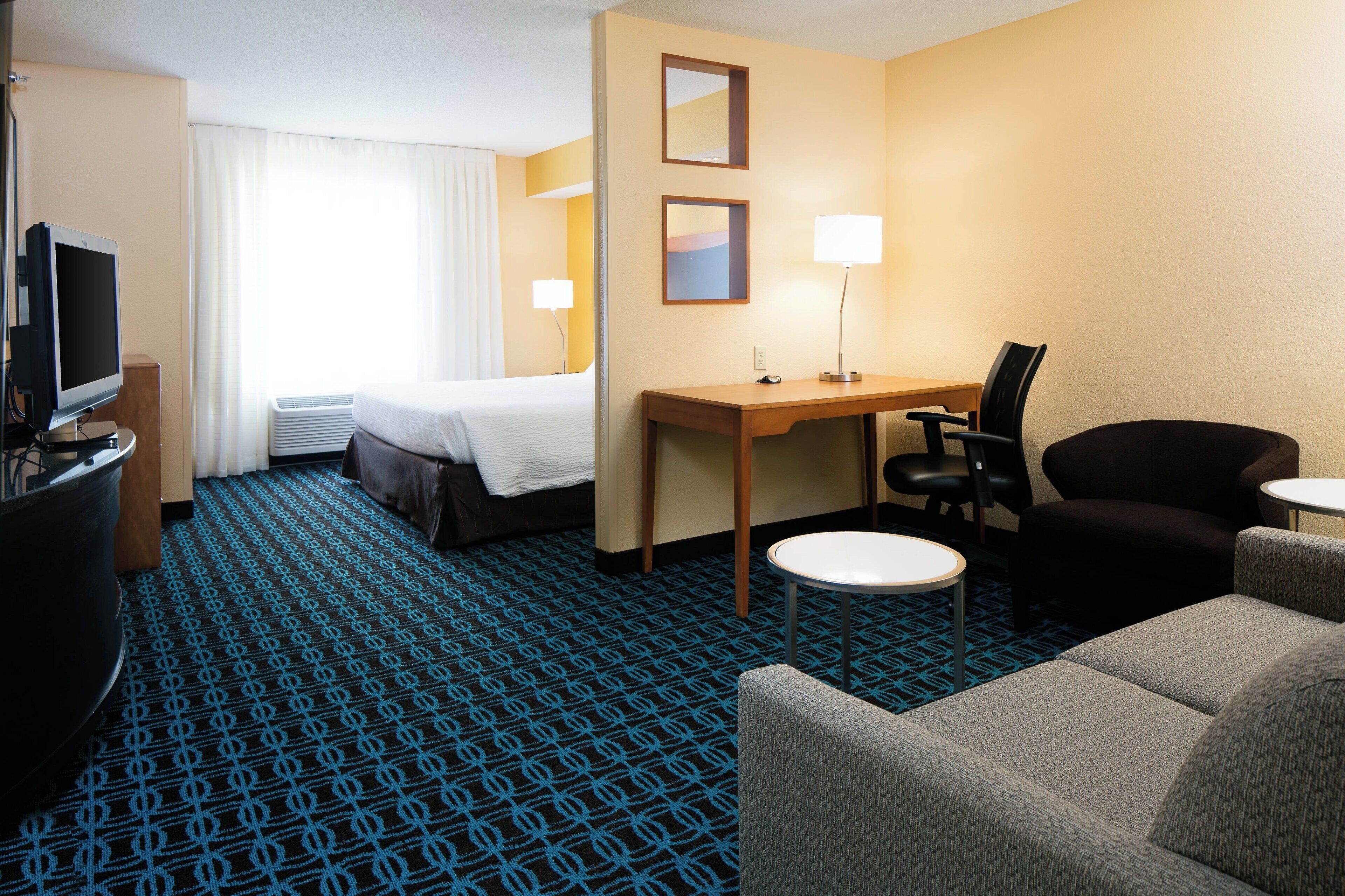 A hotel room with a bed, desk, two chairs, a TV, and a lamp.