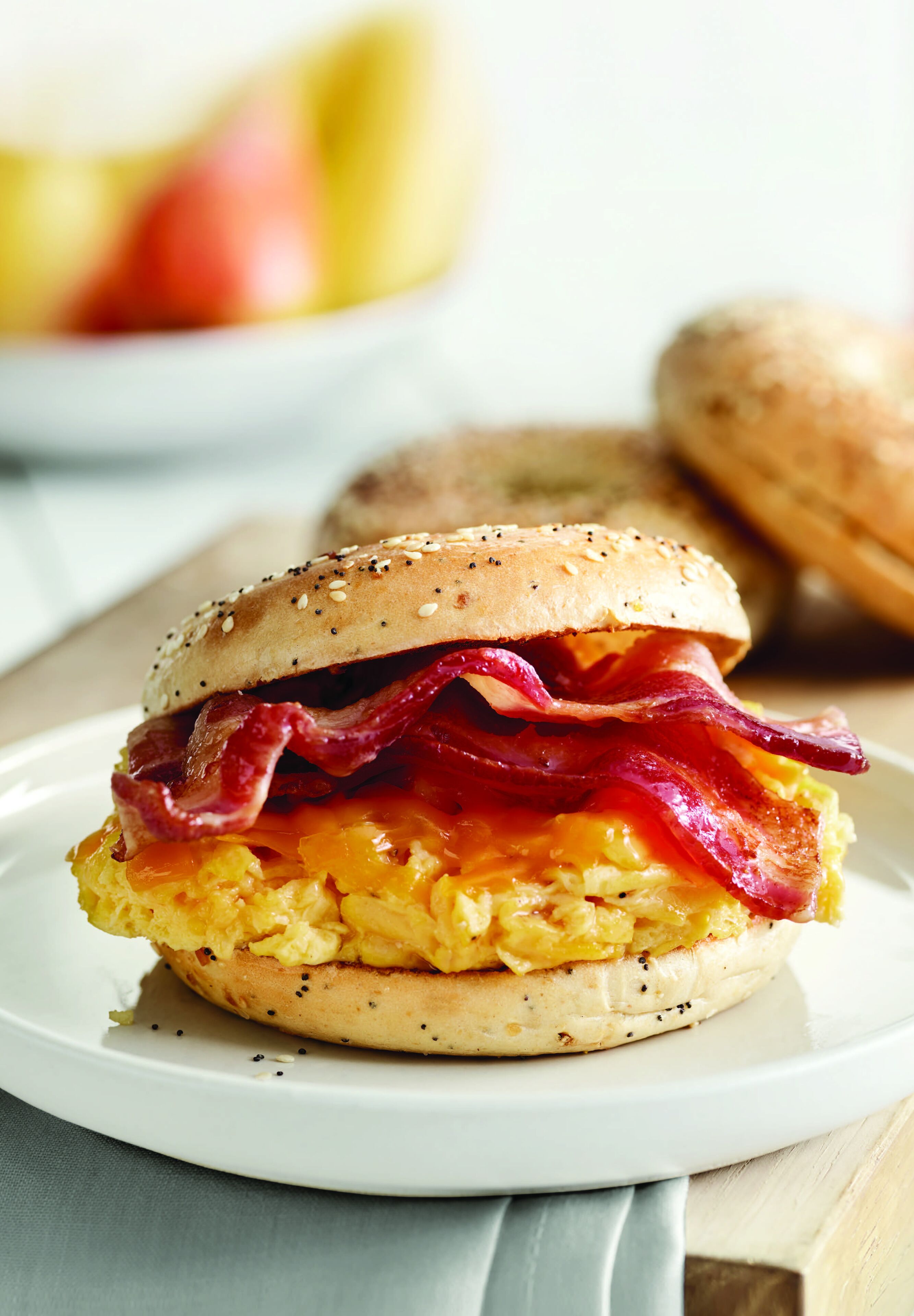 A breakfast sandwich on a white plate with a blurred background of fruits and a bagel.