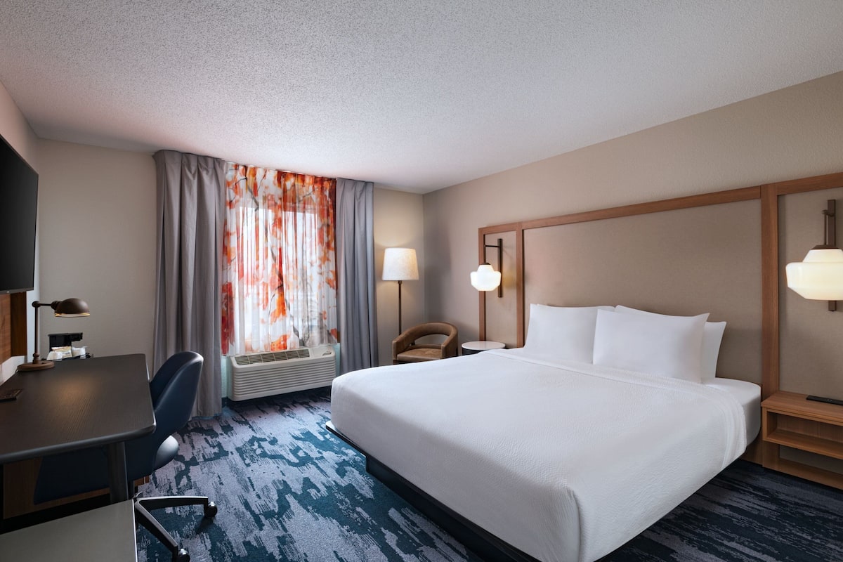 A hotel room with a large bed, a desk, a chair, a lamp, and a window with curtains.