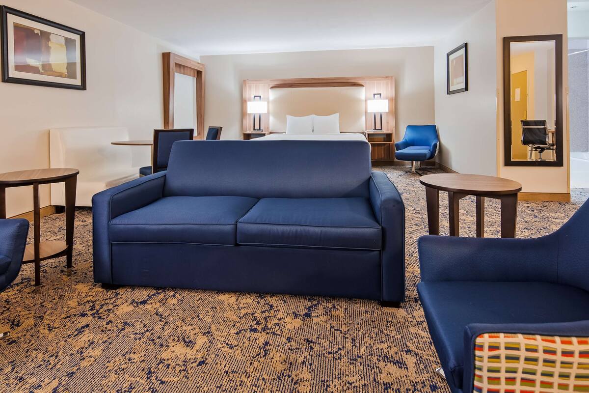 A hotel room with a large sofa, two armchairs, a small table, a bed with a wooden headboard, and a carpeted floor.