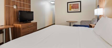 A hotel room with a bed, a TV on a wooden cabinet, a small round table, a chair, and a framed picture on the wall.