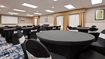 A conference room with round tables, black chairs with white sashes, a projector screen, and a large window with curtains.
