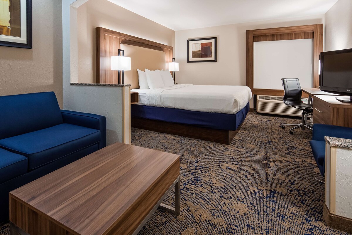 Quarto de hotel com sofá azul, mesa de madeira, cama com roupa de cama branca, escrivaninha com televisão e duas luminárias de parede.