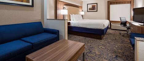 Habitación de hotel con un sofá azul, una mesa de madera, una cama con ropa de cama blanca, un escritorio con televisor y dos lámparas fijadas a la pared.