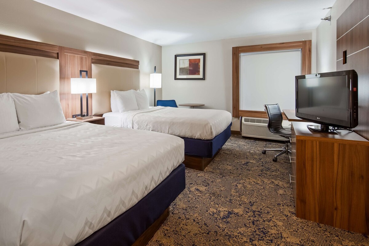 Quarto de hotel com duas camas, uma mesa com televisão, uma cadeira e um abajur.