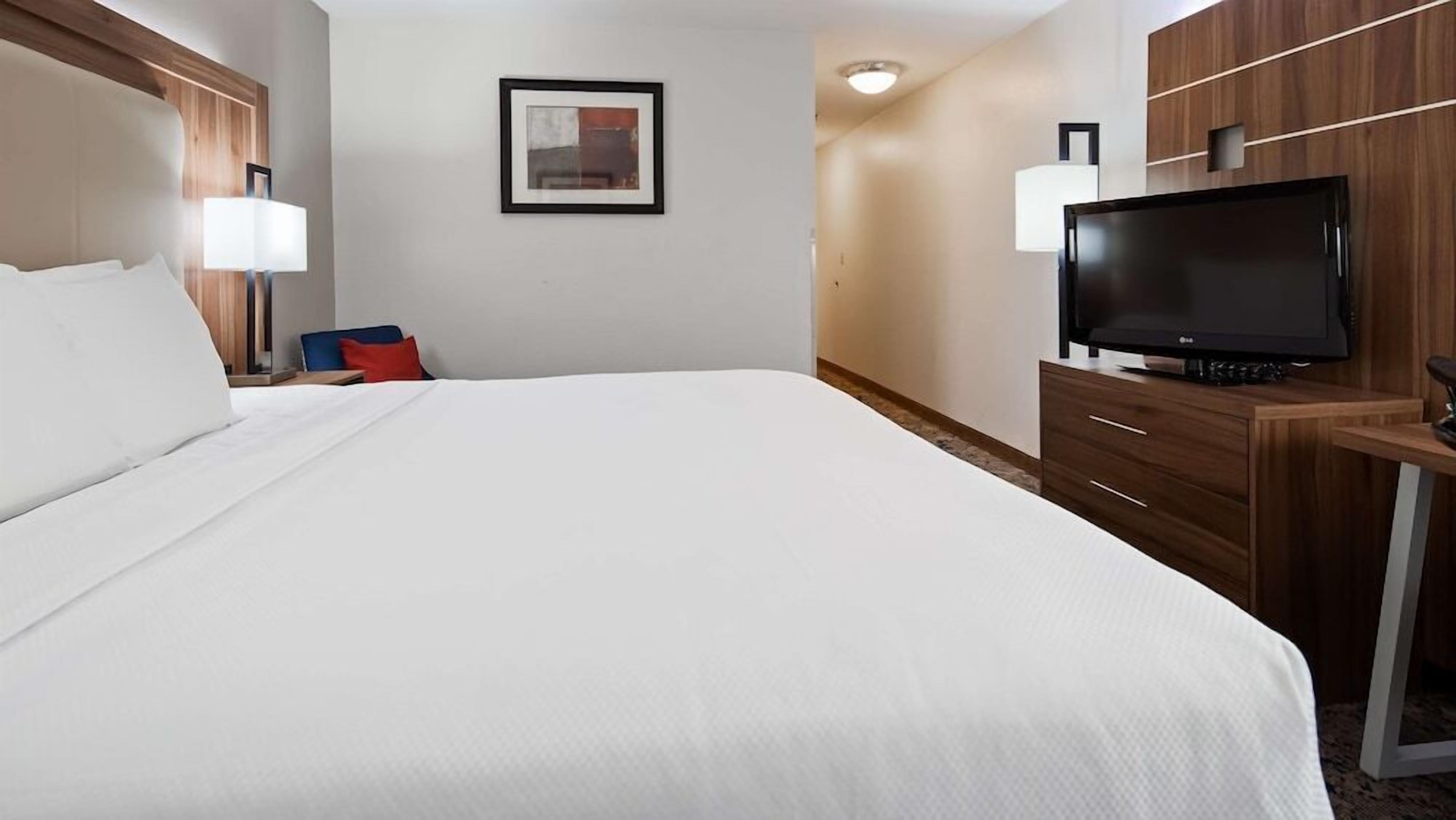 Hotellihuone, jossa on suuri sänky, työpöytä, jossa on litteänäyttöinen televisio, tuoli, yöpöytä lampun kanssa ja seinällä oleva kehystetty kuva.