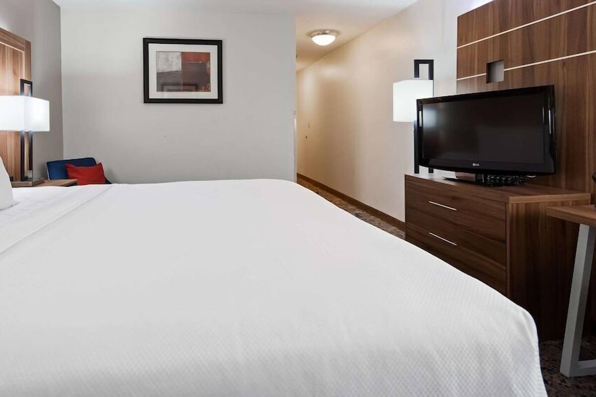 Hotellihuone, jossa on suuri sänky, työpöytä, jossa on litteänäyttöinen televisio, tuoli, yöpöytä lampun kanssa ja seinällä oleva kehystetty kuva.