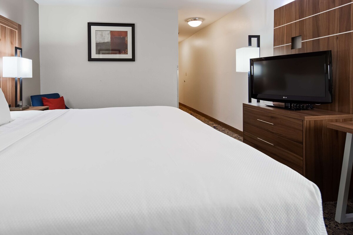 Quarto de hotel com uma cama grande, uma mesa com televisão de tela plana, uma cadeira e uma televisão fixada na parede.