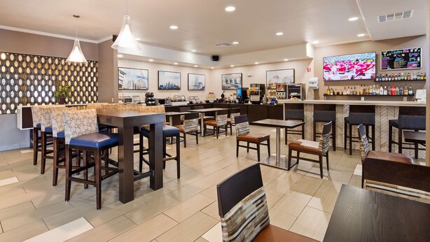 A modern restaurant with bar seating, a counter with a beverage display, and framed cityscape pictures on the walls. - Best Western Plus Kansas City Airport-KCI East (Kansas City)