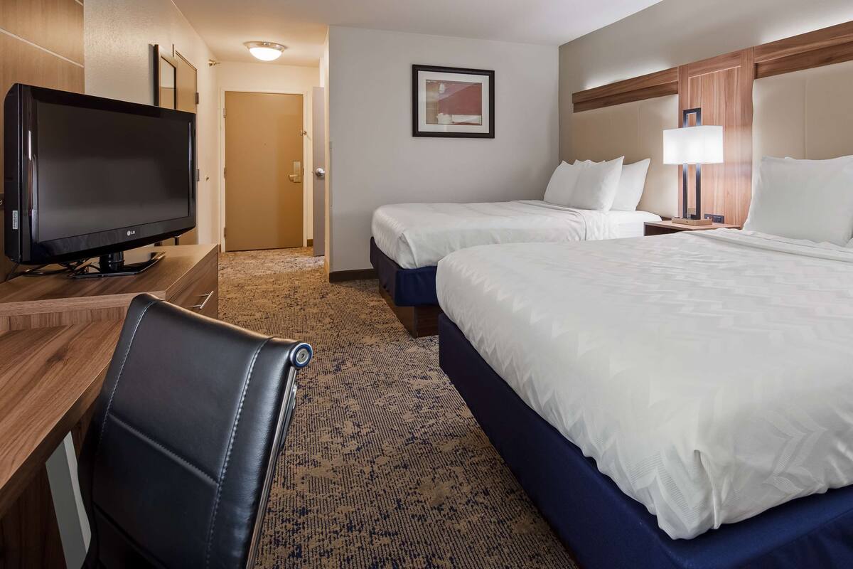 A hotel room with a large bed, a smaller bed, a desk with a flat-screen TV, a leather chair, and a carpeted floor.