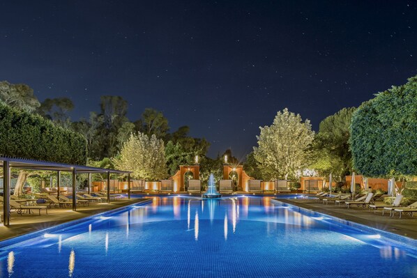 Relax by the illuminated pool surrounded by lush greenery and trees, with lounge chairs and umbrellas for a serene atmosphere.